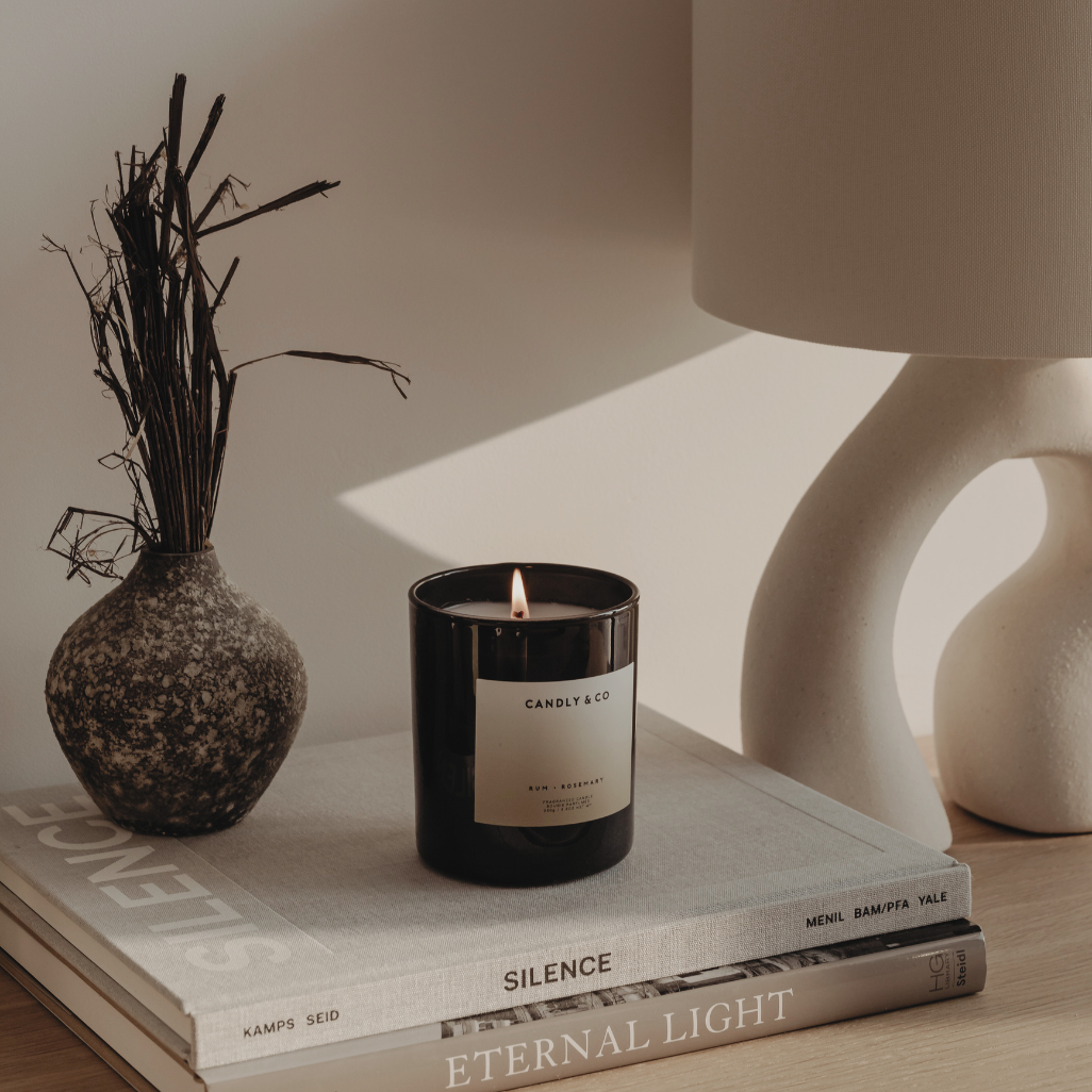 Candle with brand design showing and vase, sat on a pile of books on a desk.