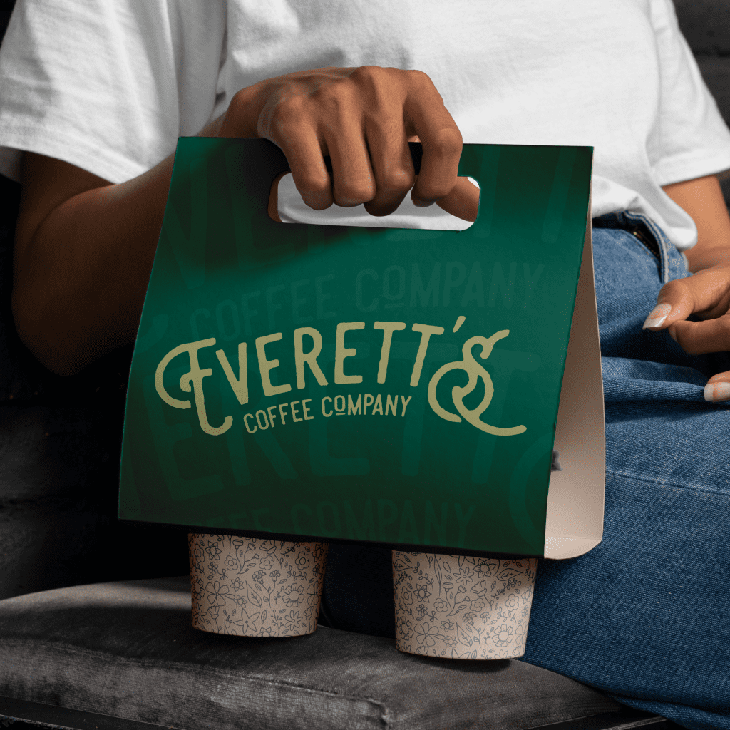 2 Coffee cups being carried in a cardboard cup holder with the coffee shop branding and the Coffee company logo in large gold lettering on green.