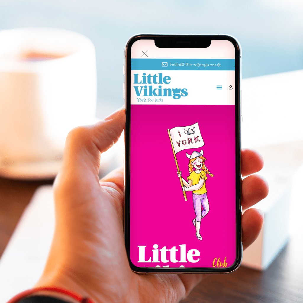 Hand holding a mobile phone that shows the home page of the Little Vikings eCommerce website with an illustration of a girl holding a flag that reads I love York